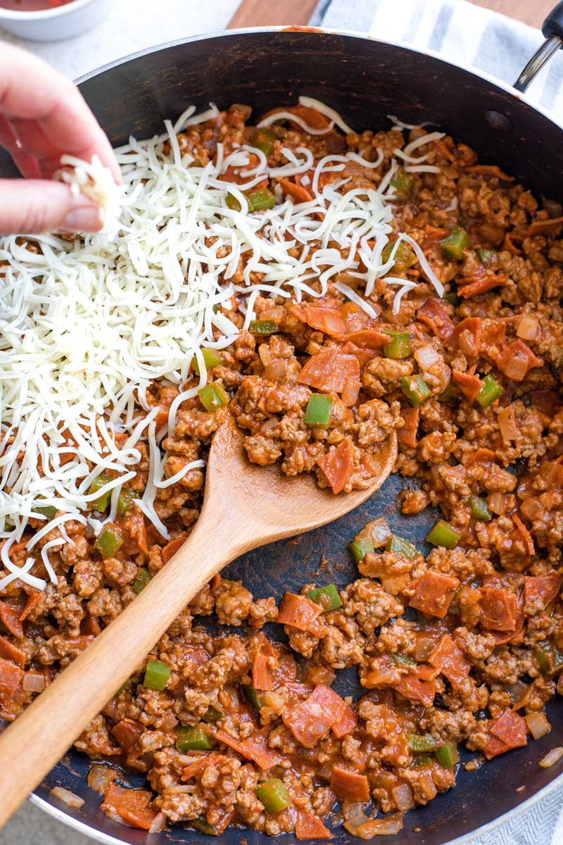 Fingers sprinkling shredded mozzarella cheese into skillet of meat.