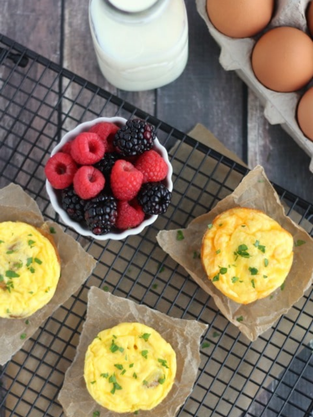 3 egg muffins on brown parchment squares on cooling rack, surrounded by bowl of berries, jug of milk and brown eggs in carton.