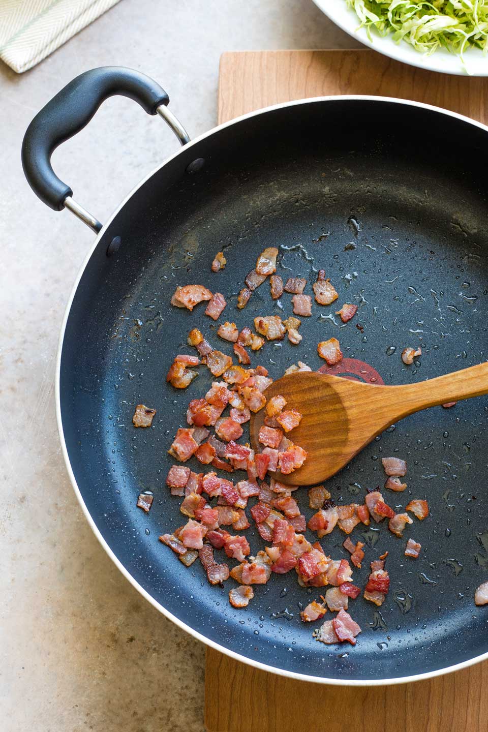 Sauteed bacon in pan being stirred with wooden spoon.
