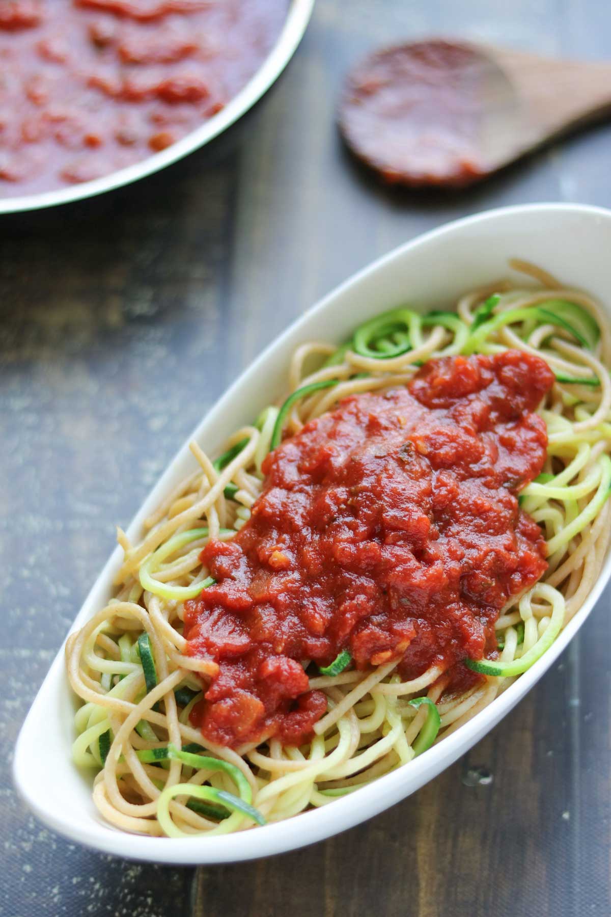 Curved white oval serving dish full full of a mix of spaghetti with zoodles, topped with meat-free marinara, with a pan of sauce and sauce-covered wooden spoon in background.