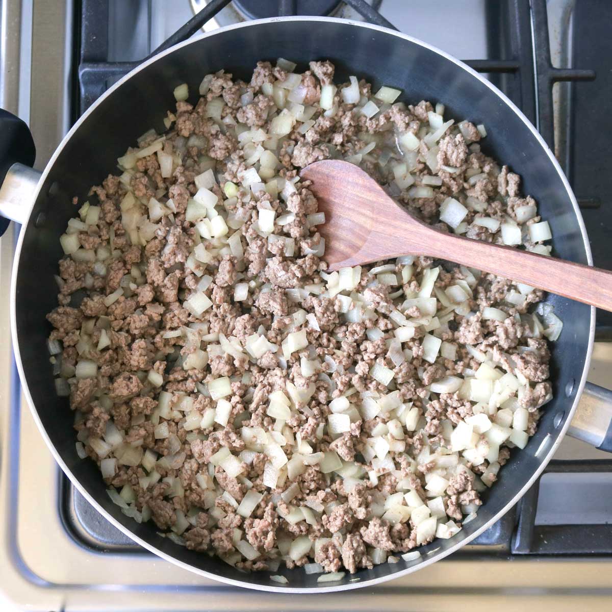 Overhead of large skillet on stove with cooked onions and ground beef with wooden spoon stirring.
