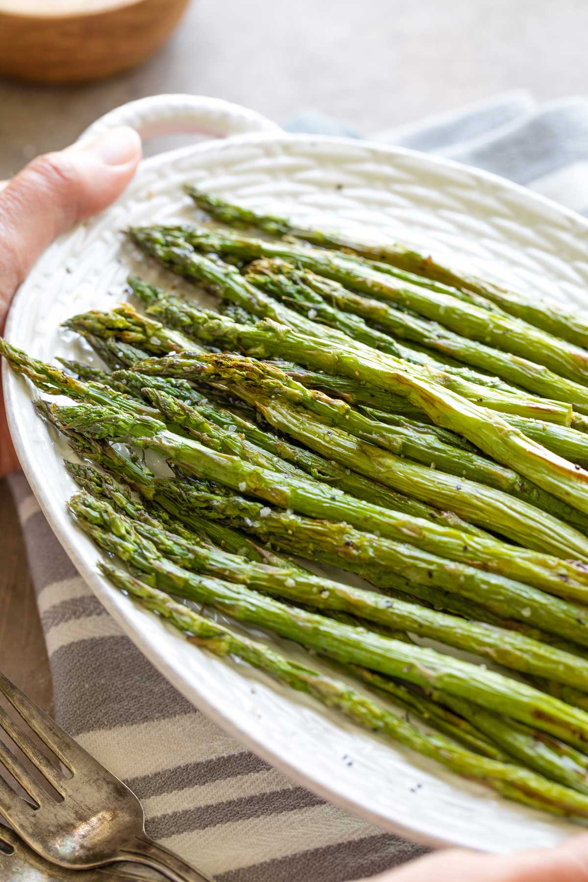 Hands holding serving platter of asparagus above gray-striped cloth.