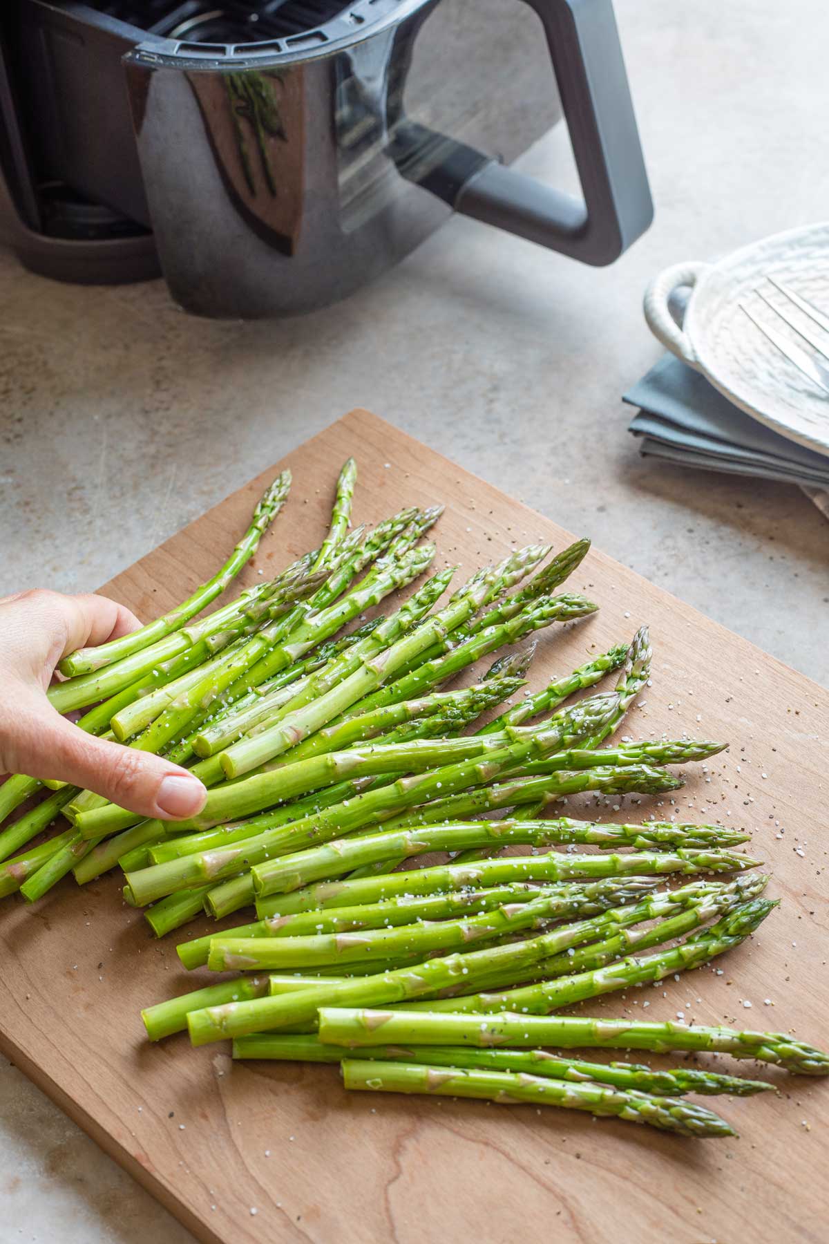 Hand reaching in to toss raw asparagus with extra virgin olive oil and seasoning before being put into the air fryer in background.