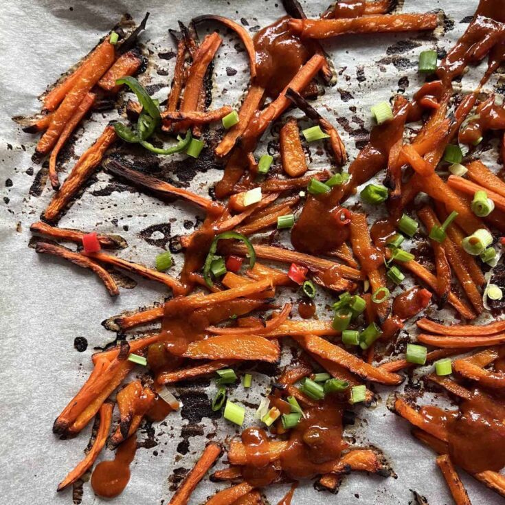 Butternut squash fries on white parchment after baking, drizzled with sauce and garnished with sliced green onions.