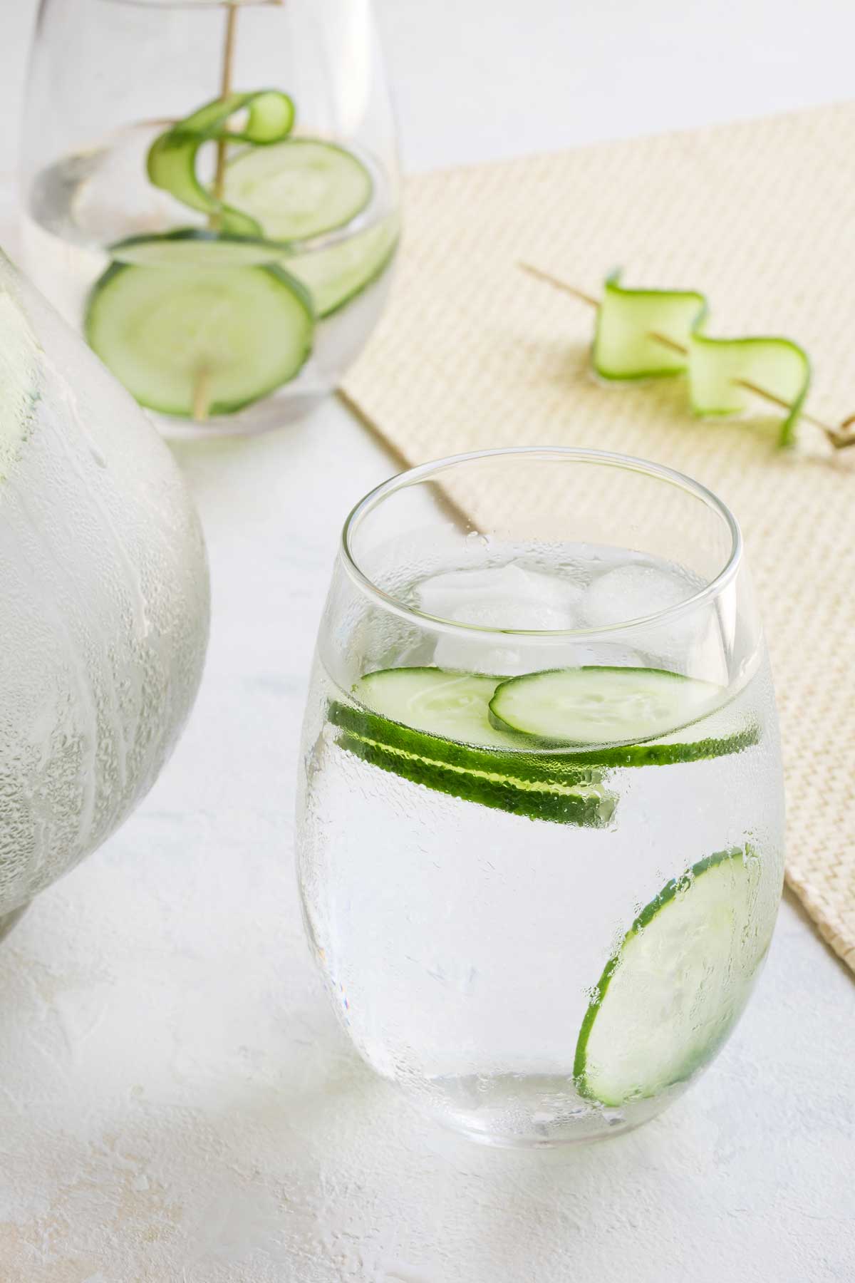 Side closeup of one water glass with four cucumber slices and a few ice cubes floating, plus a second glass and pitcher in background.