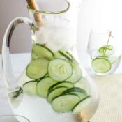 Side view of finished cucumber water recipe served in a pretty glass pitcher, with filled glasses and a decorative skewer at edges of photo.