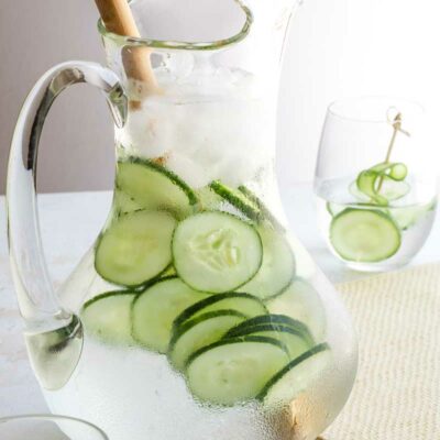 Full pitcher of water with cucumber slices and ice, and a wooden spoon for stirring, with two glasses nearby.