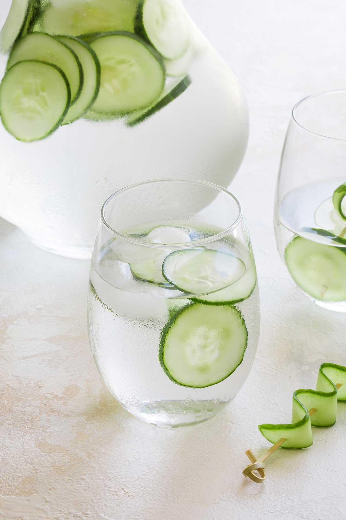 One glass of iced cucumber water in foreground, surrounded by a second glass, a filled pitcher, and a decorative garnish.