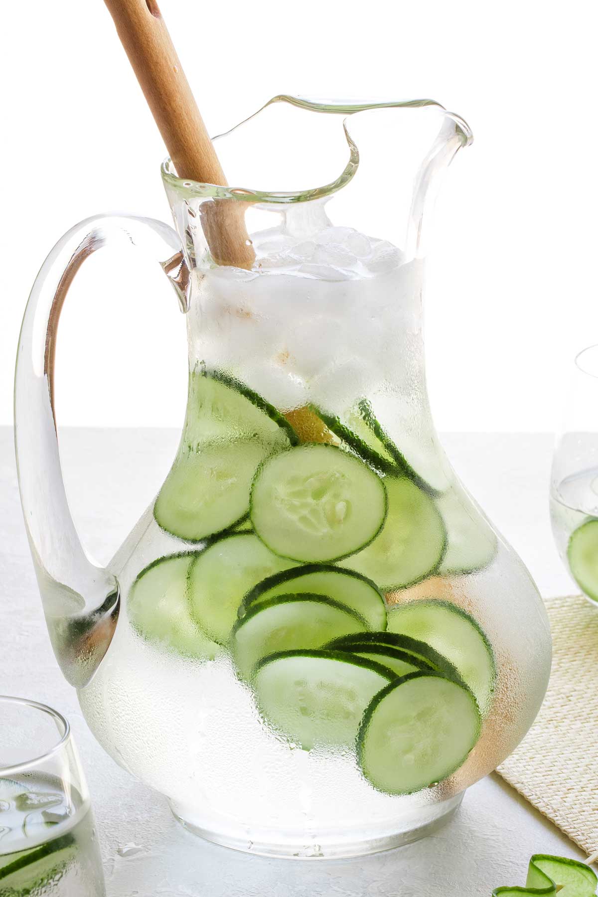 Side view of a glass pitcher of water filled with cucumber slices, topped with ice, with a wooden stirring spoon in it.