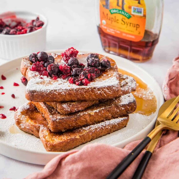 Four slices stacked on white plate, topped with berry compote with utensils, pink napkin and syrup bottle.