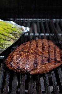 Steak on grill grates with seared grill marks, and asparagus on tray in background.