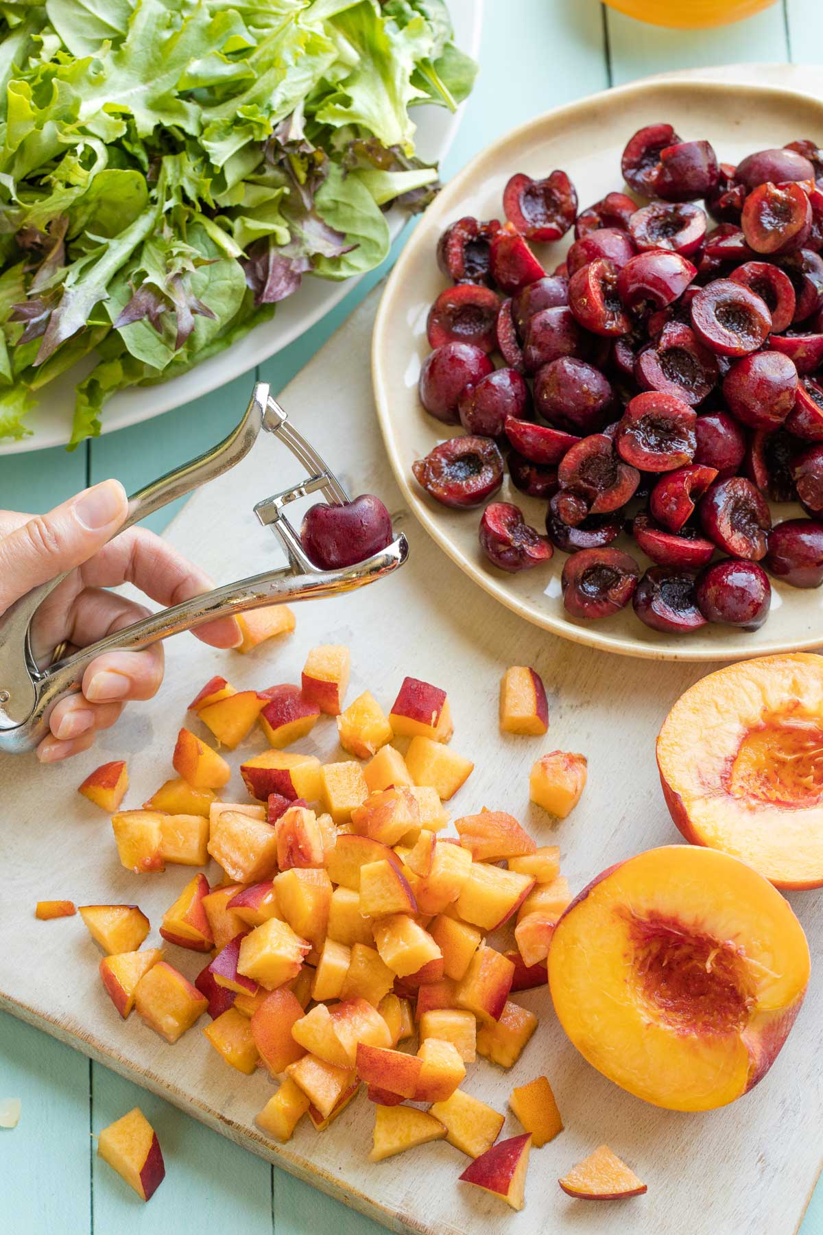 Peaches being cut into cubes on white cutting board with one peach halved, and plate of halved cherries with hand using cherry pitter.