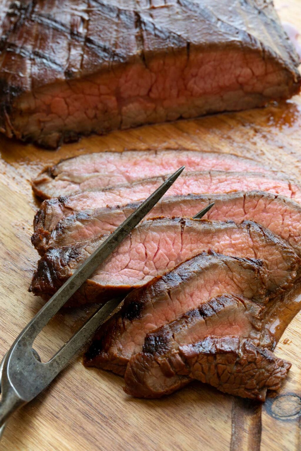 Tines of antique carving fork twined in several slices of steak on wooden board.