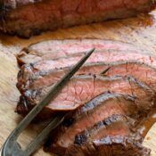 Tines of antique carving fork twined in several slices of steak on wooden board.