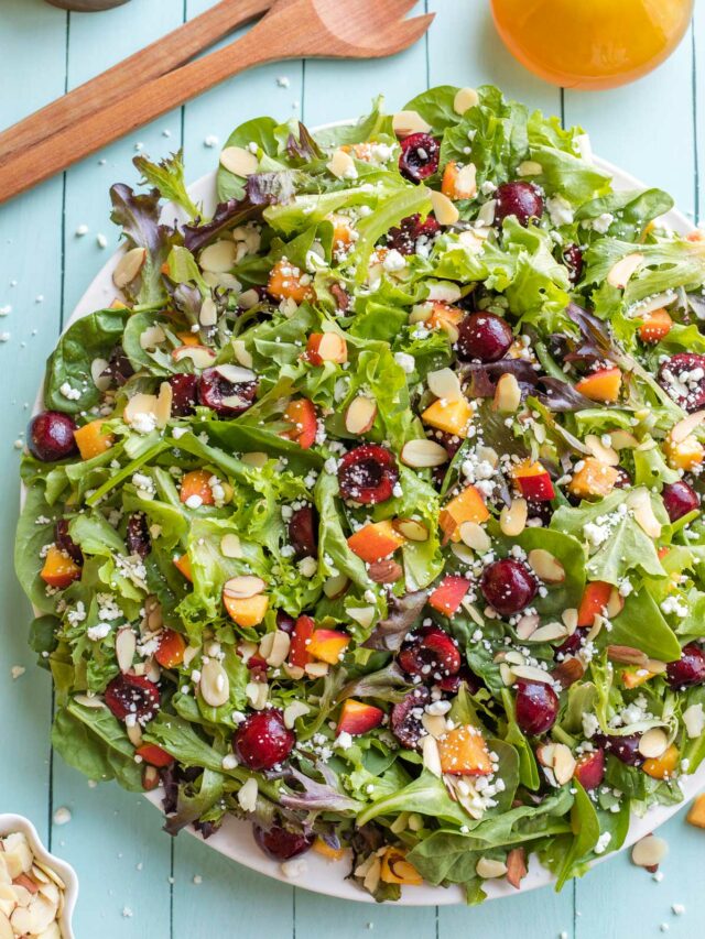 Overhead of finished peach salad with extra nuts, goat cheese, dressing and wooden salad set.