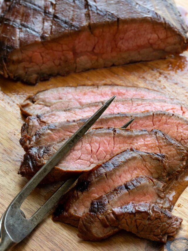 Tines of antique carving fork twined in several slices of steak on wooden board.