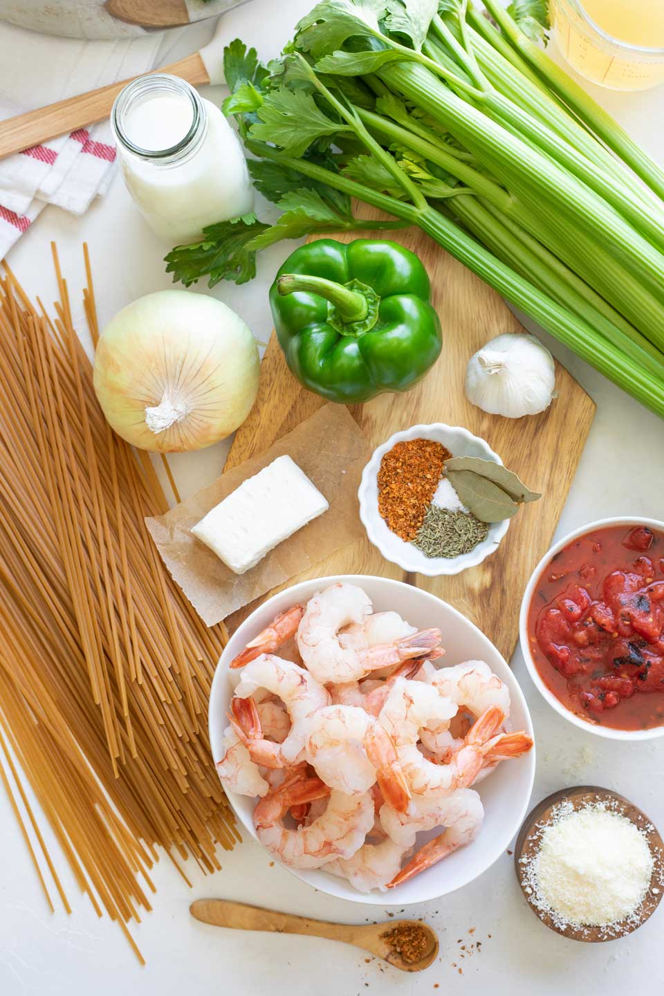 Overhead of ingredients, including raw shrimp in bowl, Cajun spices, dry pasta, and uncut veggies gathered on wooden cutting board.