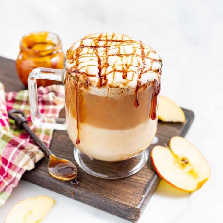 One drink in glass mug on wooden board, dripping caramel with apples and jar of caramel nearby.