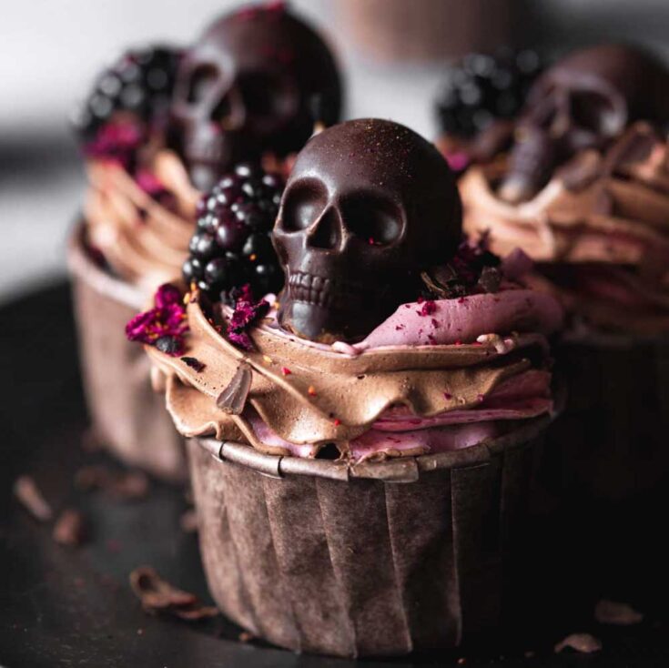 Closeup of one finished cupcake, decorated with vegan frosting, a chocolate Halloween skull and a blackberry, with others in background.