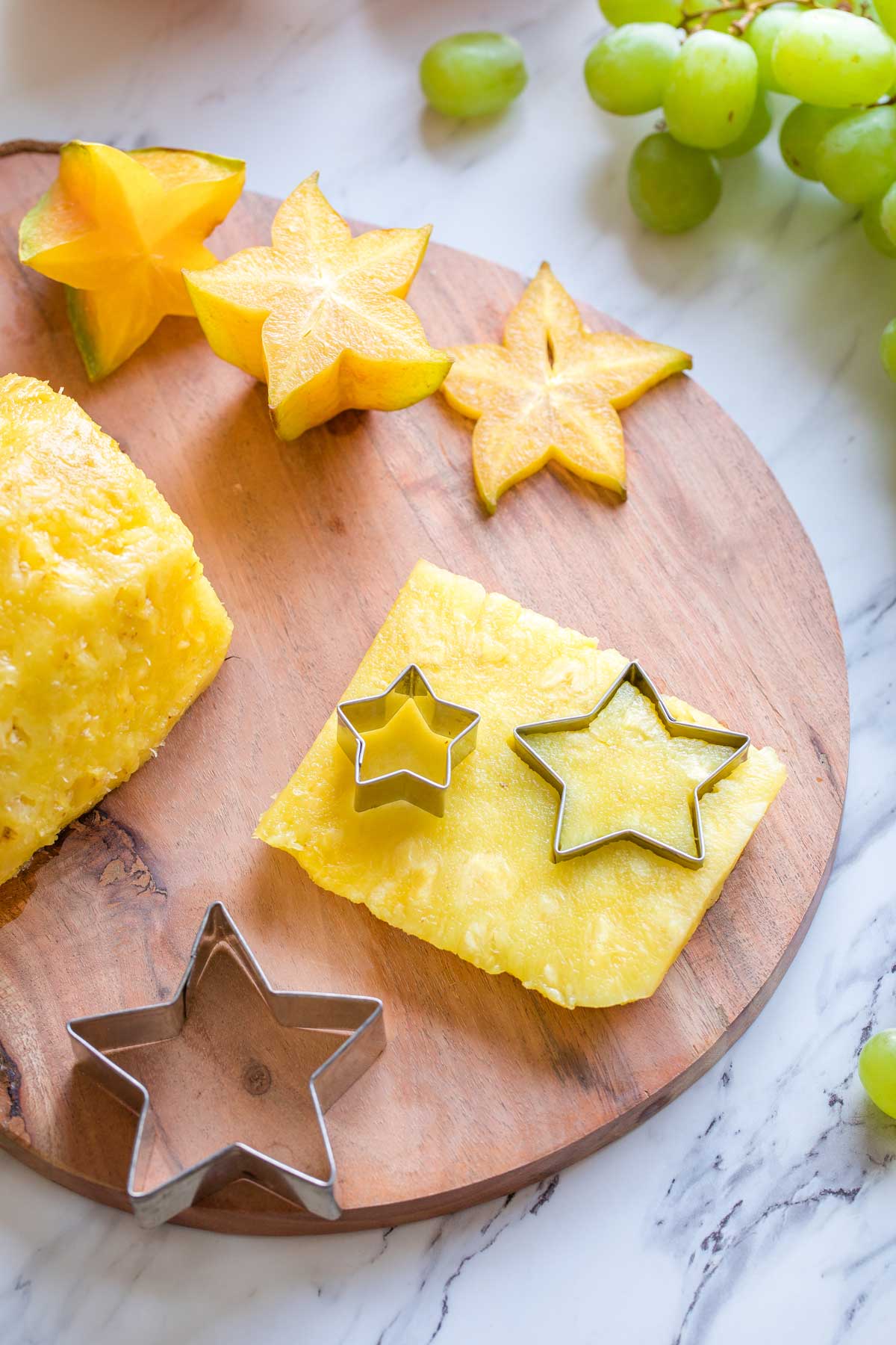 Star-shaped cookie cutters making stars from a plank of pineapple, with starfruit and green grapes alongside.