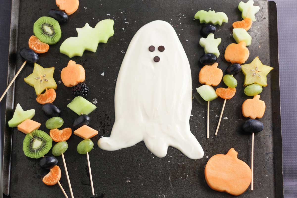 7 kabobs and fruit cutouts of a pumpkin and a bat, arranged on a black tray around white, ghost-shaped fruit dip.