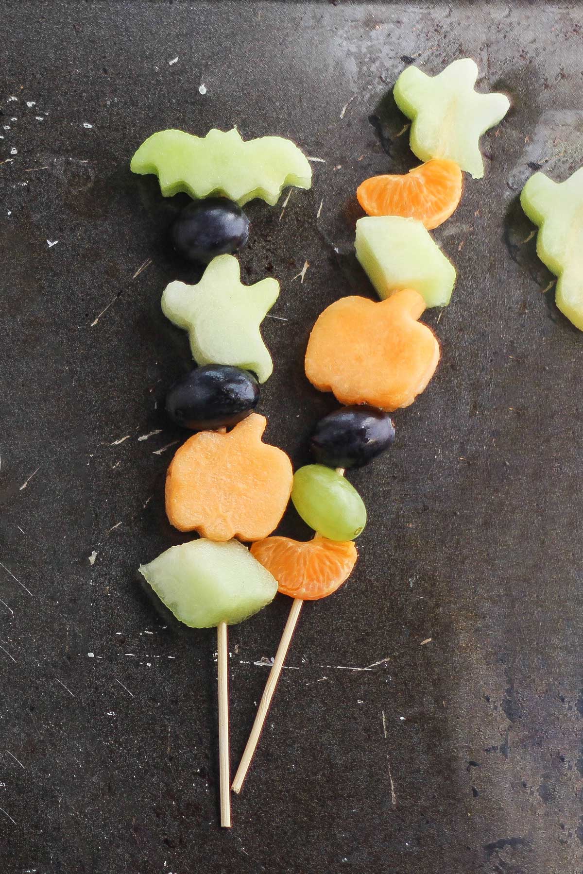 2 kabobs with fruit chunks and also cutout fruit shapes of a bat, pumpkins and ghosts.