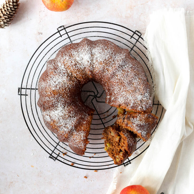 Overhead flatlay of cake with several slices cut out, on a round cooking rack with a pinecone and apples nearby.