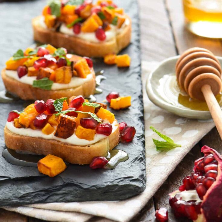 Three crostini on slate serving tray with pomegranate and honey dipper alongside.