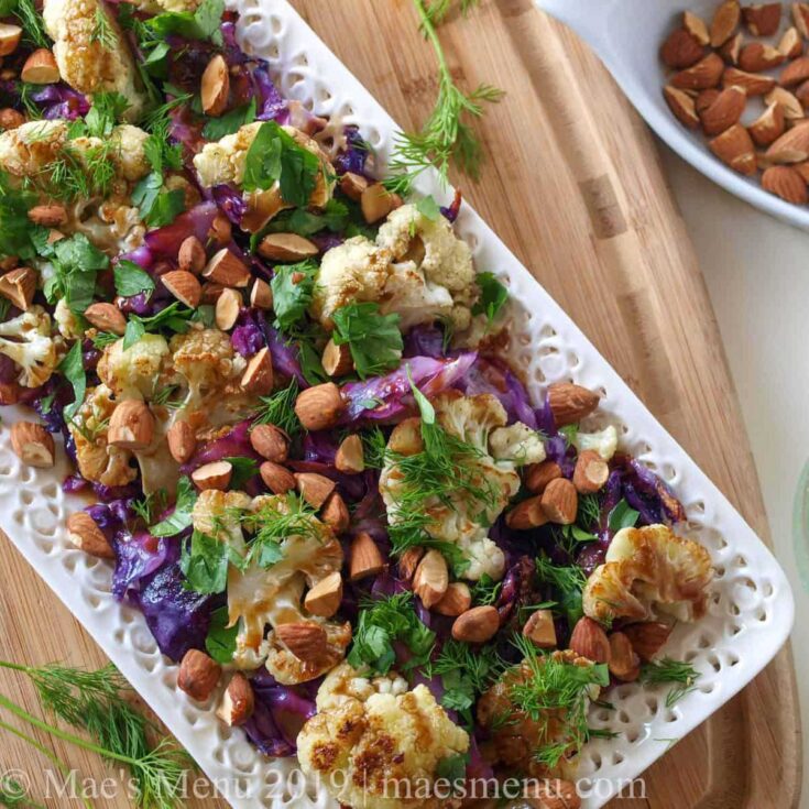Finished salad on decorative white rectangular platter with bowl of nuts nearby.