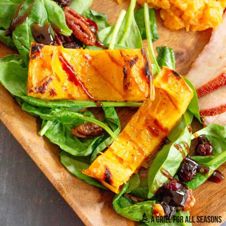 Closeup of cut squash amid greens, cranberries and nuts on wooden plate next to other Thanksgiving foods like turkey and mashed sweet potatoes.