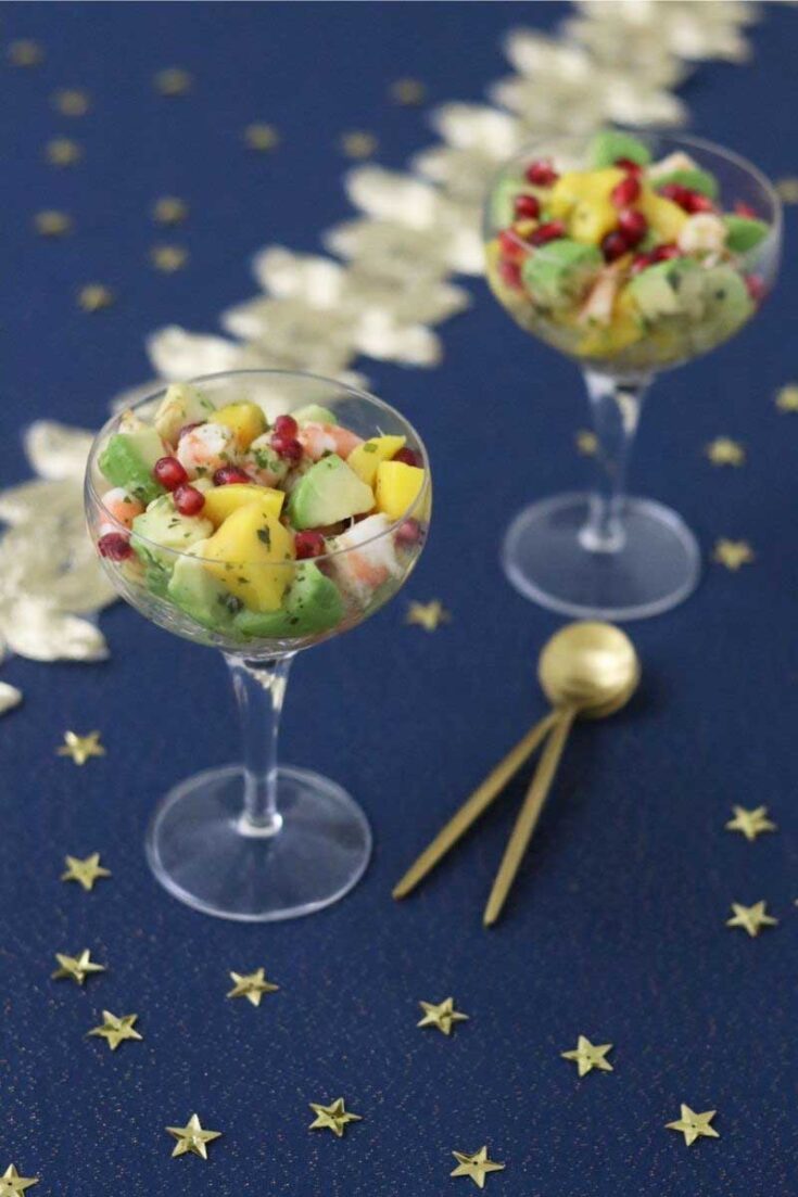 Two wine glasses full of shrimp appetizer on blue cloth decorated with stars and two golden spoons.