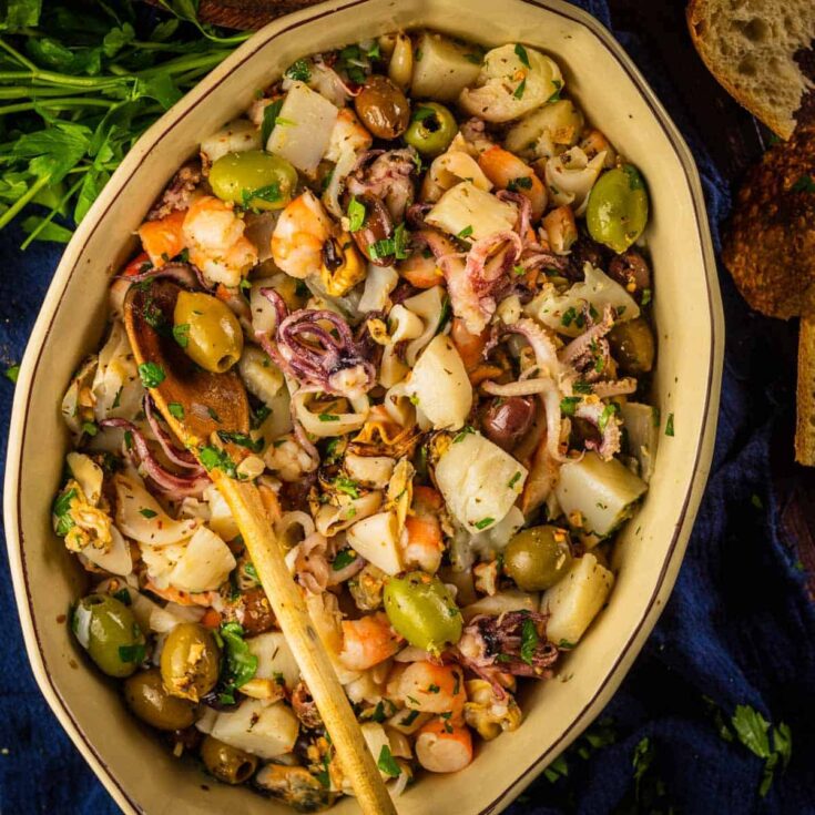 Overhead of salad in tan oval server with wooden spoon, surrounded by parsley stems and bread pieces.