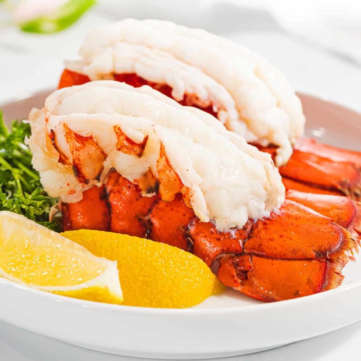 Side closeup of two lobster tails in white dish with lemon wedges.