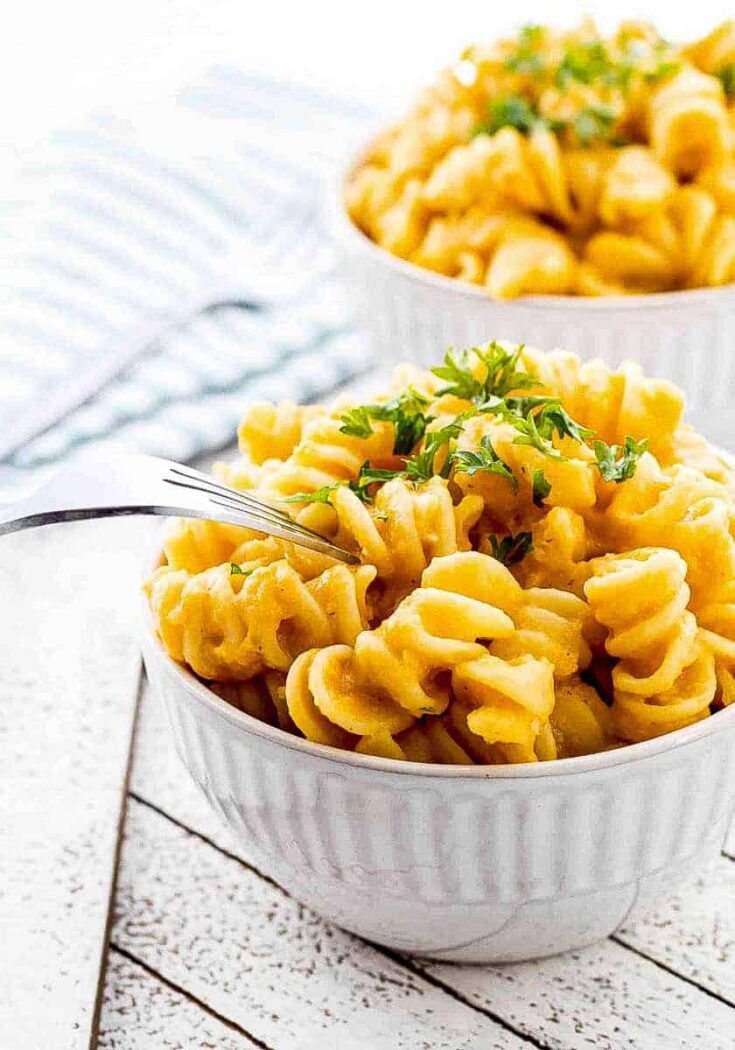 Closeup of mac and cheese heaped into decorative small white bowl, garnished with parsley with fork tines poking in and a second bowl in background.
