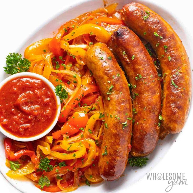 Serving platter with 3 sausages, peppers and small white dish of sauce.