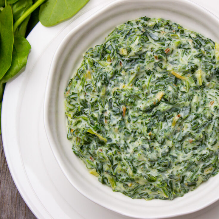 Overhead of creamed spinach in white serving bowl on white platter, with a few uncooked spinach leaves at corner of photo.