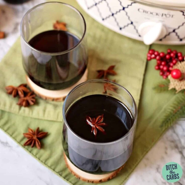 Two stemless glasses of wine on woodcut coasters atop green napkins with star anise, crockpot and decorative Christmas berries alongside.