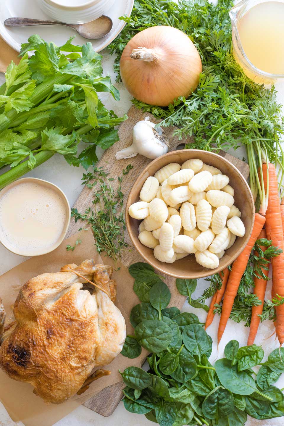 Soup ingredients: whole rotisserie chicken, bowl of frozen gnocchi, onion, head of garlic, carrots with tops on, spinach leaves, chicken broth, fresh thyme and milk.