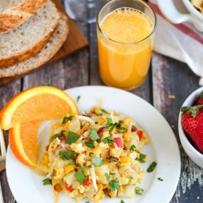 Pinnable image of one serving on white plate with oranges, strawberries, OJ, bread and serving bowlful of breakfast scramble surrounding.