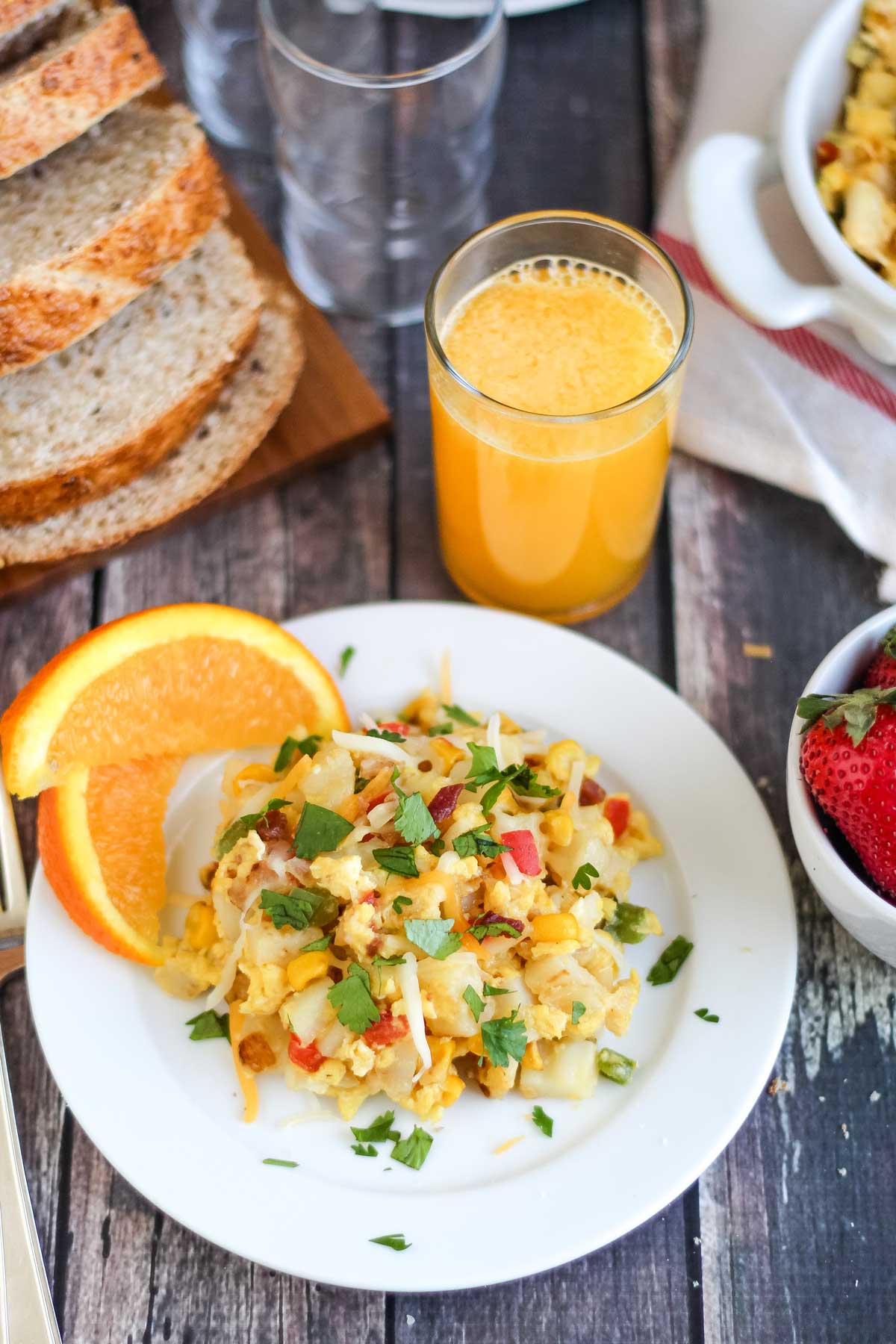 Breakfast scene featuring skillet scramble at center on white plate and in background in shite serving bowl, along with sliced oranges, glass of orange juice, little bowl of fresh strawberries, and wheat bread.