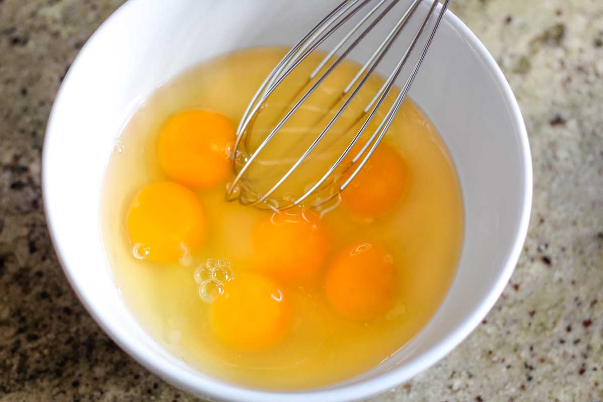 White bowl with 6 raw eggs, with wire whisk ready to begin lightly beating them.