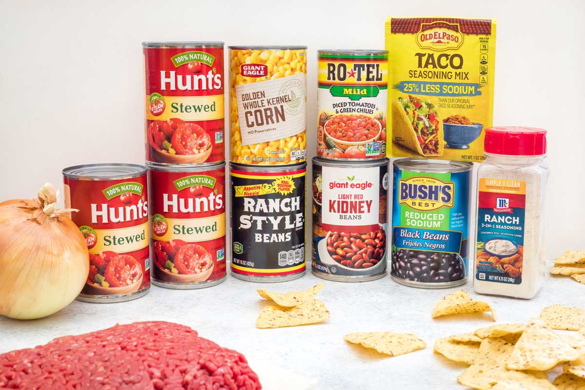 Unopened cans and containers of soup ingredients, stacked two-high, with ground beef, unpeeled onion and taco chips.