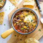 Closeup flatlay of one bowl of taco soup surrounded by whole chips, spoon and second bowlful.