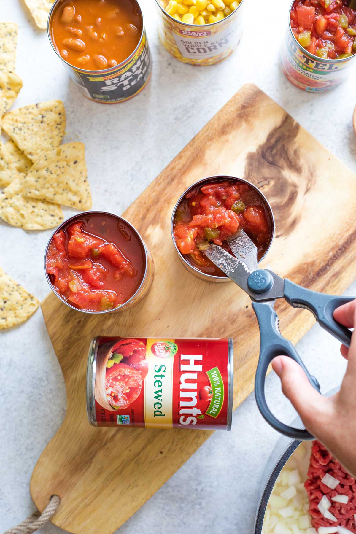 Hand using kitchen scissors to chop tomatoes still in can, with other cans of ingredients, soup pot and taco chips surrounding.
