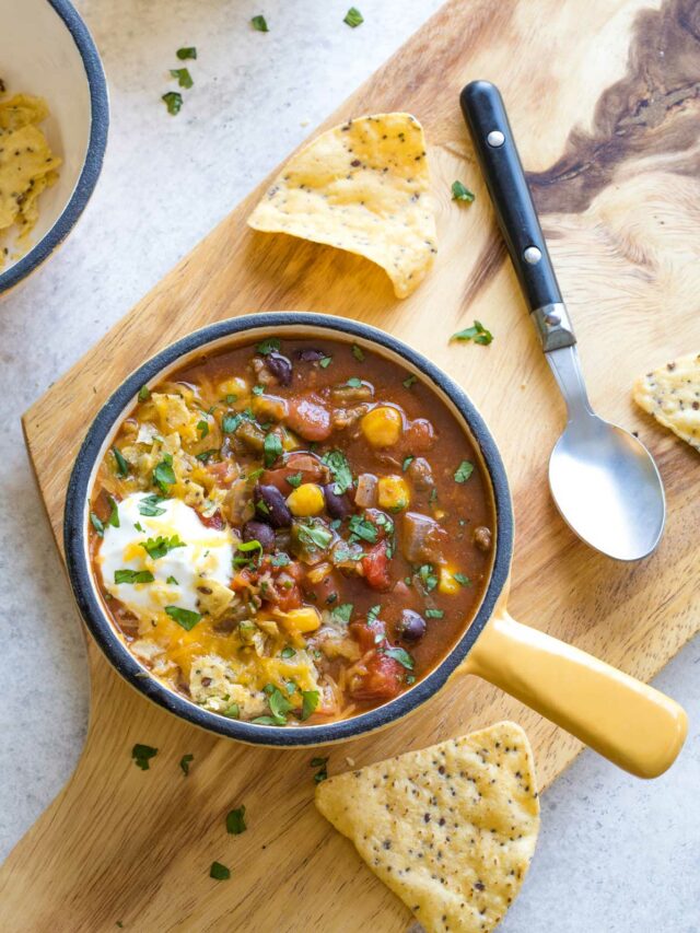 Overhead of 1 handled bowl of soup on wooden board with spoon, taco chips, and garnishes nearby.