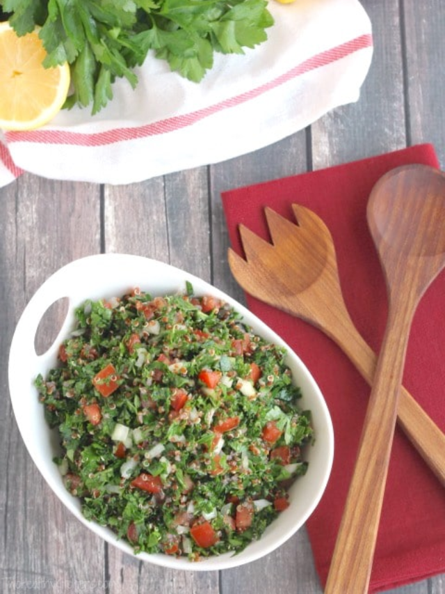 Scene with quinoa tabouli in serving bowl, wooden servers on burgundy napkin, and extra ingredients nearby.