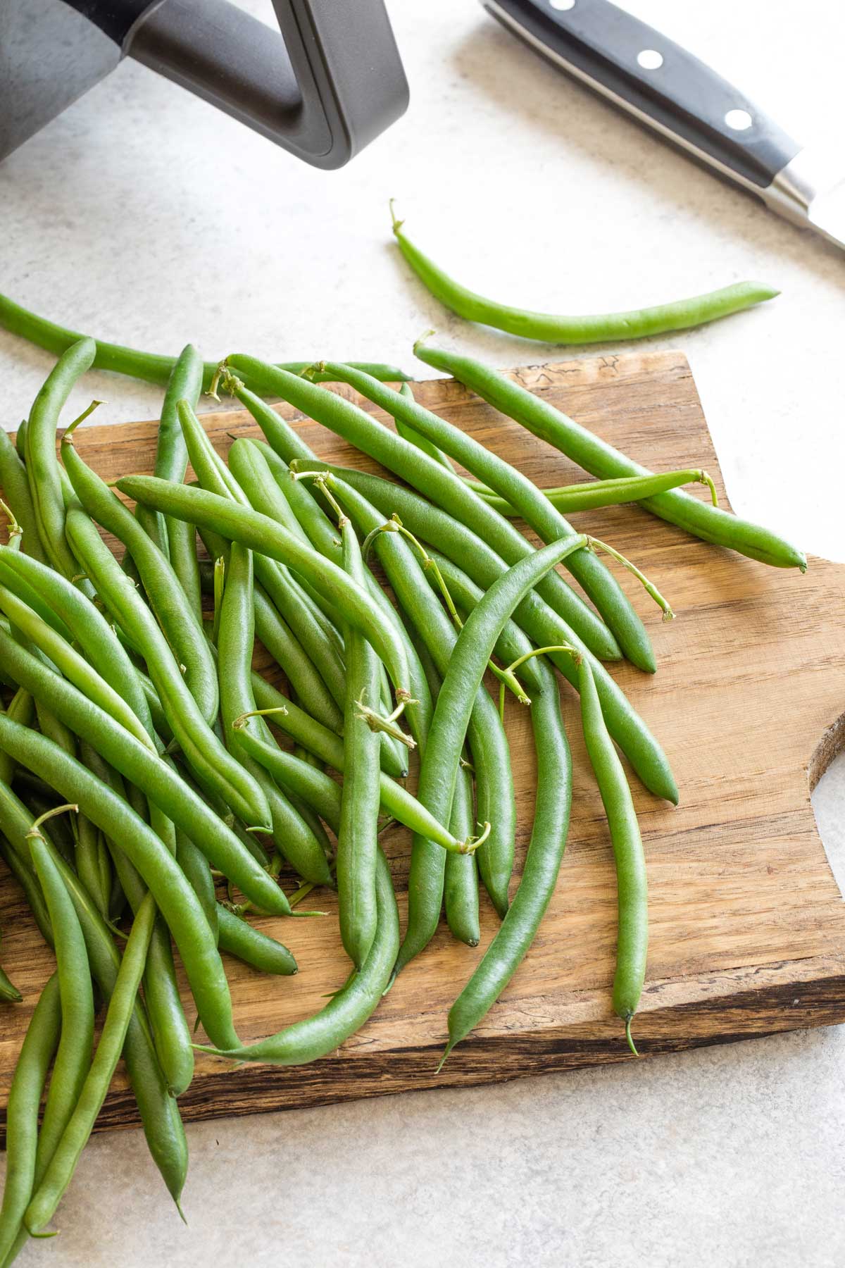 Raw, fresh green beans tumbled across wooden cutting board with air fryer in background.