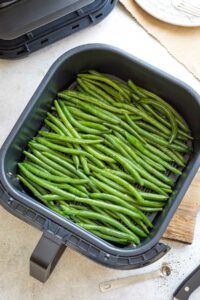 Overhead of raw, fresh green beans spread out in air fryer basket before cooking.