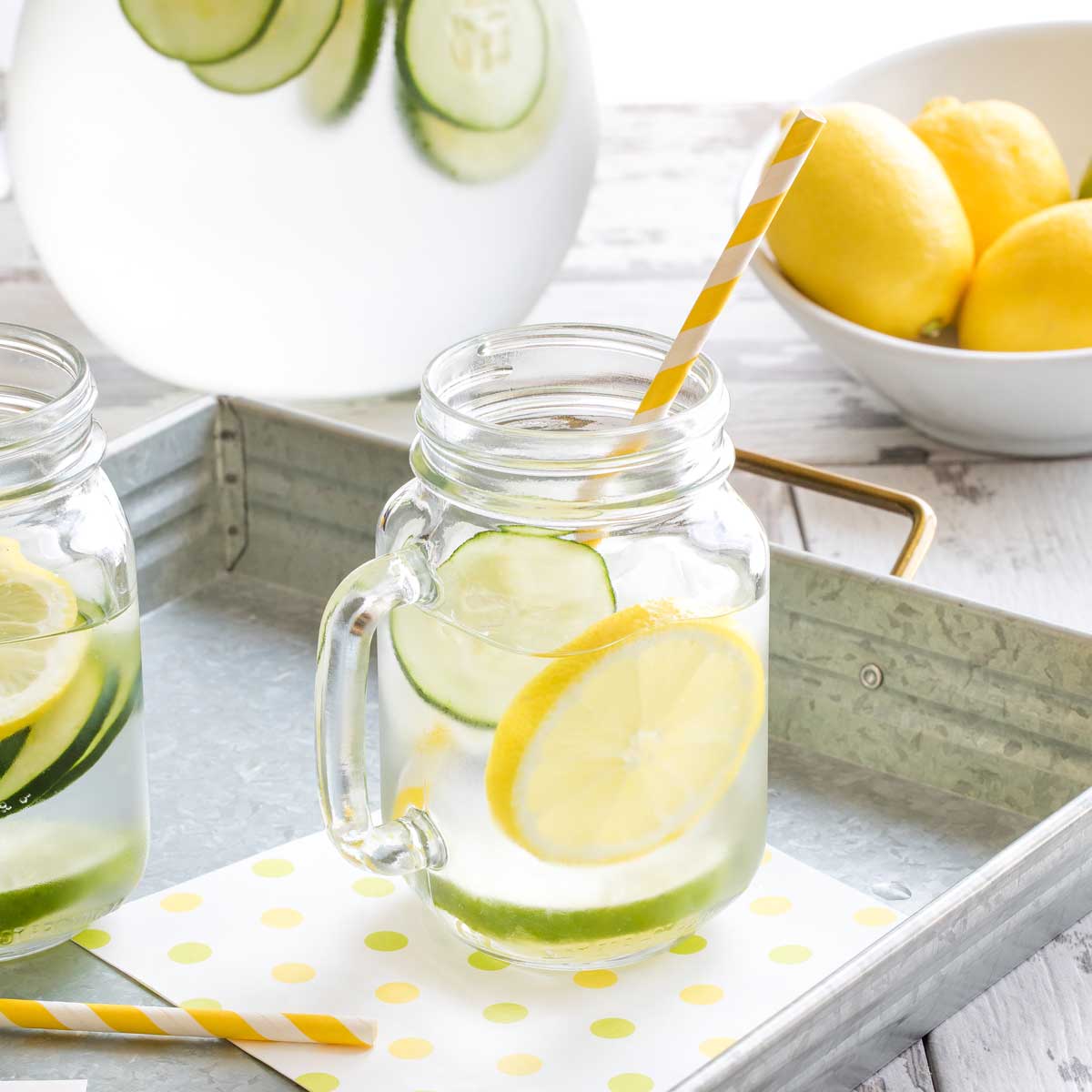 Closeup of one clear handled glass of water with a slice of lemon, cucumber and lime and a yellow striped straw, with glass pitcher of water and bowl of whole lemons in background.