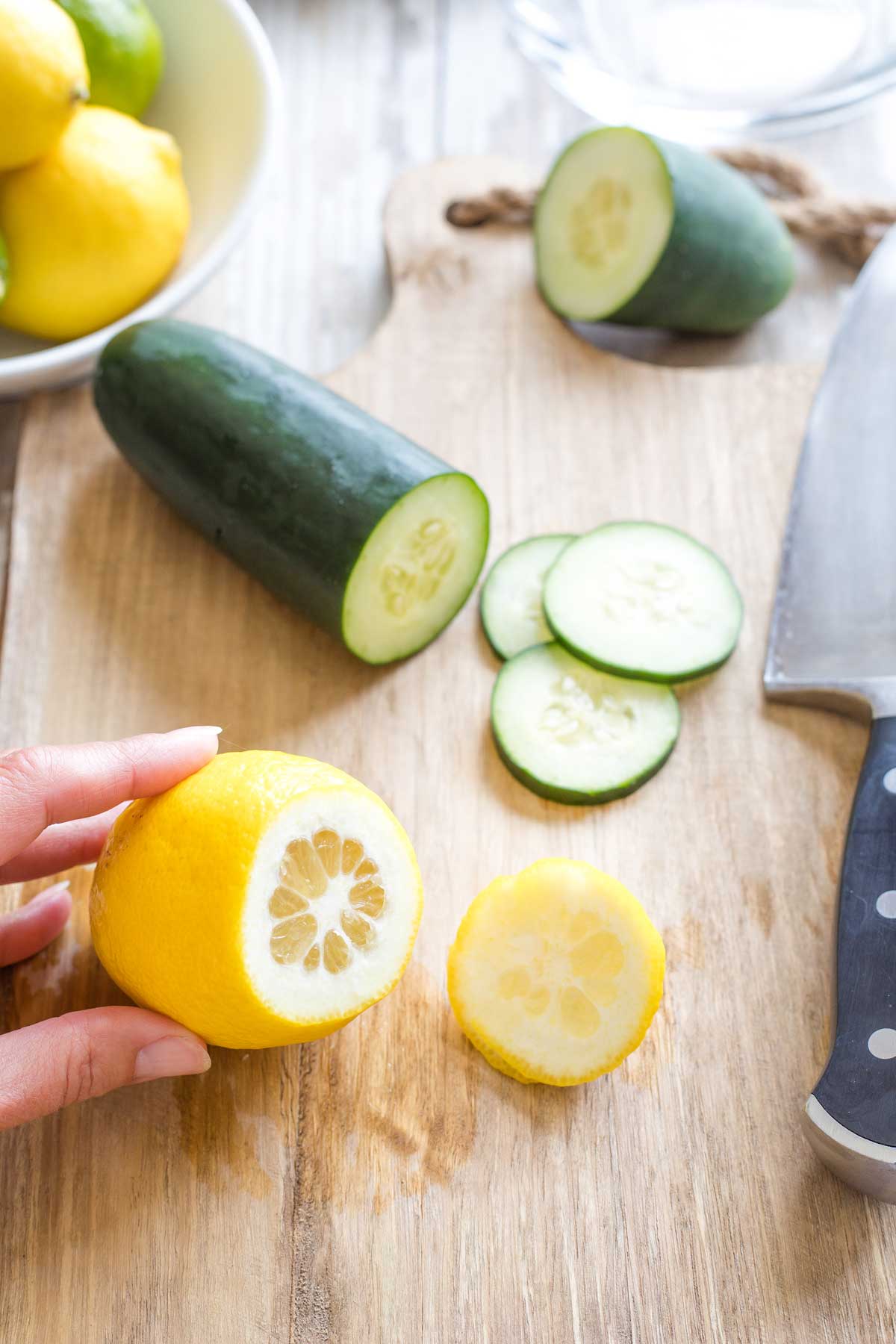 Hand holding lemon with end cut off on cutting board; partially sliced cucumber and extra lemons and limes in background.